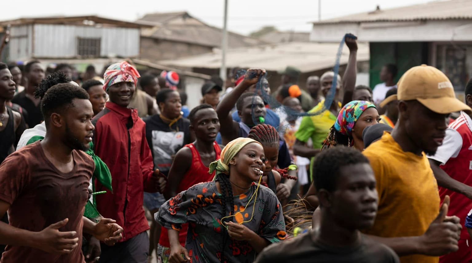 A crowd of people moves along a street in daylight, some smiling, some with fists raised.