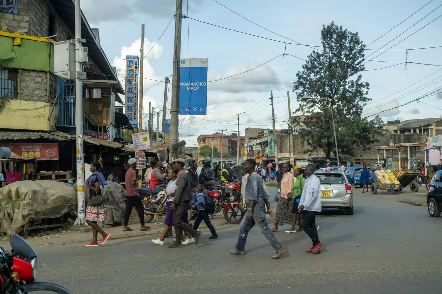 People crossing a busy street in a Nairobi neighbourhood, with shopfronts, motorbikes and a billboard behind them.