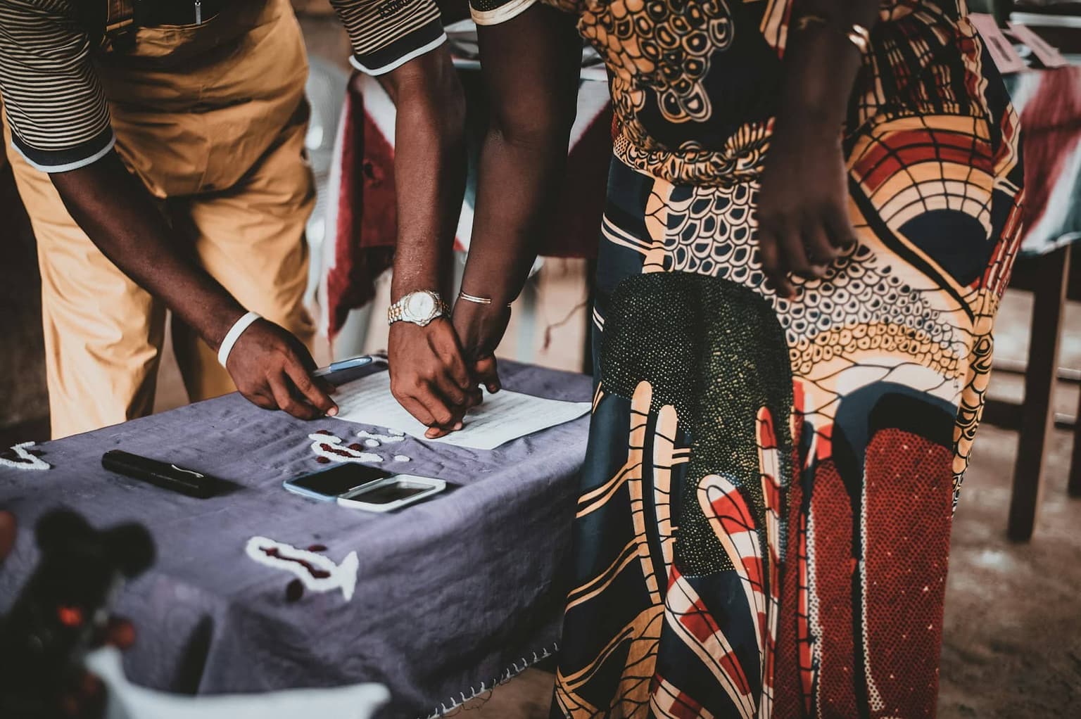 Close-up of a voter pressing an inked thumb onto a paper register on a cloth-covered table, an official's hands beside it.